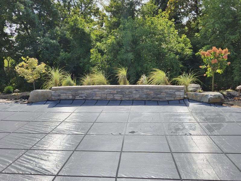 Rectangular black pool cover in the foreground, stone retaining wall with ornamental grasses and small trees in the background, surrounded by green trees.